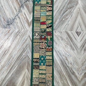 May include: A rectangular, patchwork table runner with a dark green border. The runner features a variety of patterned fabric squares in shades of green, gold, red, and black. The design includes floral and geometric motifs.