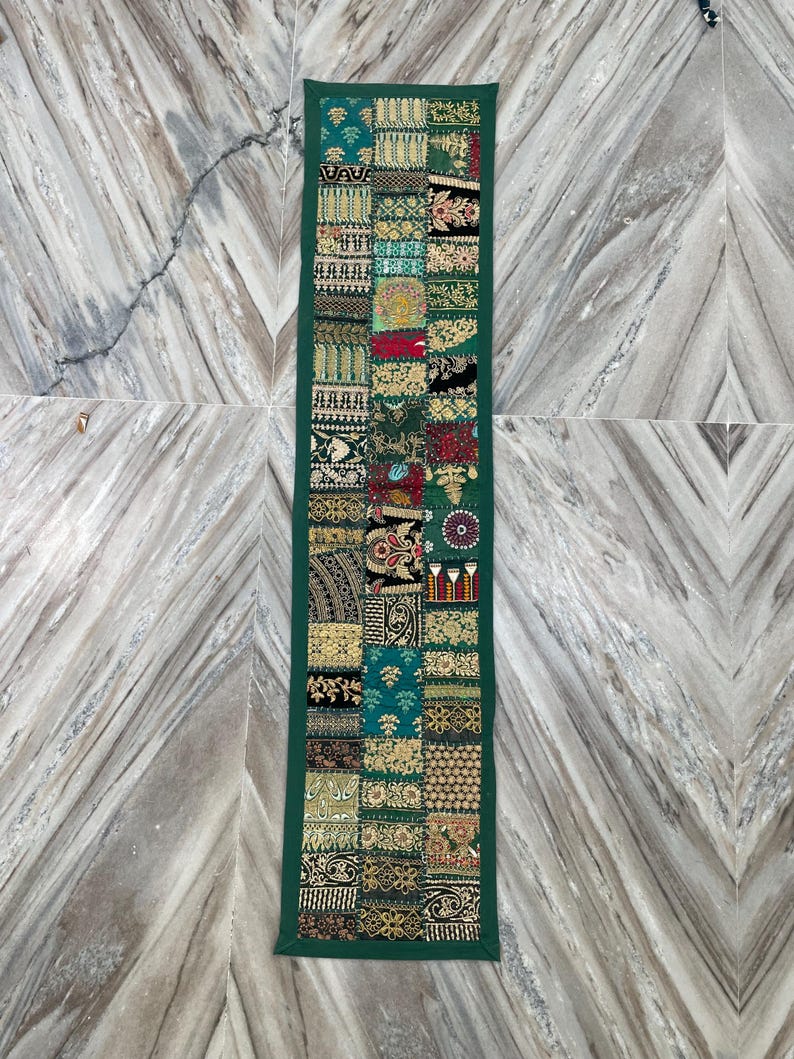 May include: A rectangular, patchwork table runner with a dark green border. The runner features a variety of patterned fabric squares in shades of green, gold, red, and black. The design includes floral and geometric motifs.
