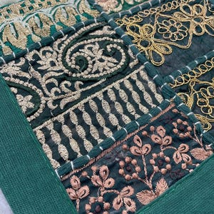 May include: Close-up of a patchwork textile with intricate embroidery. The design features squares of dark green fabric with gold and rose-colored floral and geometric patterns. A solid green border frames the design.