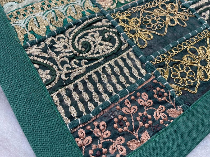 May include: Close-up of a patchwork textile with intricate embroidery. The design features squares of dark green fabric with gold and rose-colored floral and geometric patterns. A solid green border frames the design.