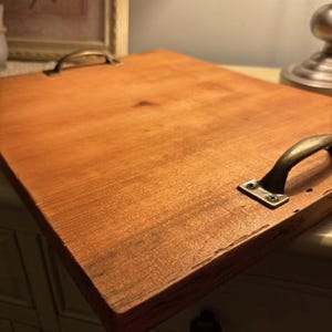 May include: A rectangular wooden serving tray with a warm, reddish-brown stain. It features two antique brass handles and is designed for carrying items. The tray is likely handmade and suitable for home decor.