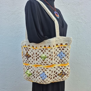 Pode incluir: Bolsa tote de crochê creme com um padrão floral. A bolsa apresenta um design de grade com motivos florais coloridos em verde, azul e amarelo. Possui alças resistentes e um interior espaçoso, perfeito para transportar o essencial diário.