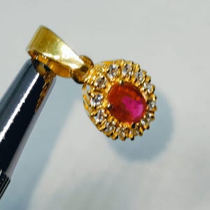 May include: A gold pendant with a ruby center stone, surrounded by small clear gemstones. The pendant is held by a silver tool against a white background. The pendant's bail is also gold.