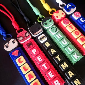 May include: A collection of five vertical superhero-themed keychains with colourful nylon cords. Each features a character head, a symbol, and block letters spelling names including CLARK, PETER, BATMAN, HULK, and a partial name on the blue keychain.