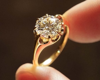 Op de afbeelding: Een gouden ring met een grote, sprankelende diamant. De diamant is rond geslepen en gezet in een gedetailleerde, sierlijke gouden zetting. De ring wordt tussen twee vingers gehouden, wat het ontwerp en de schittering benadrukt.
