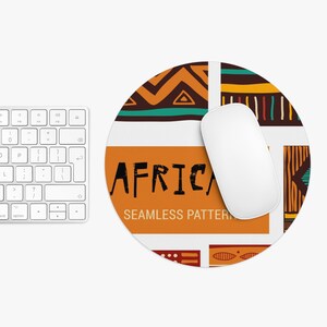 Puede incluir: Alfombrilla de ratón redonda con un diseño de inspiración africana. La alfombrilla presenta un fondo blanco con patrones geométricos en naranja, marrón y verde azulado. La palabra "AFRICA" está impresa en negro sobre un rectángulo naranja. Un ratón de ordenador blanco descansa sobre la alfombrilla.