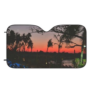 Pode incluir: Para-sol de carro com uma cena de pôr do sol tropical com palmeiras em silhueta contra um céu laranja e vermelho vibrante. O design inclui um corpo de água e um primeiro plano escuro.