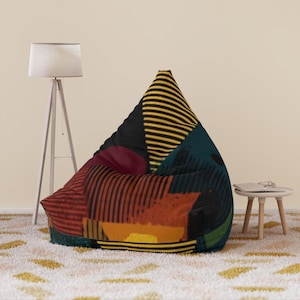 May include: A large, triangular beanbag chair with a colourful geometric pattern. The design features stripes, rectangles, and triangles in shades of black, yellow, red, orange, and teal. The beanbag is on a patterned rug.