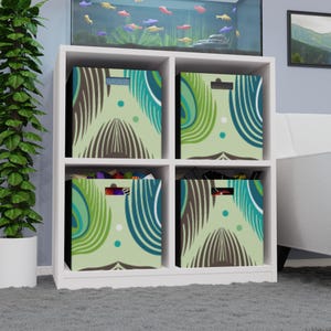 Puede incluir: Un mueble de almacenamiento cúbico blanco con cuatro cubículos, cada uno con un contenedor de almacenamiento estampado. Los contenedores presentan un diseño con formas de plumas turquesas, verdes y marrones sobre un fondo verde claro. La unidad está sobre una alfombra gris.