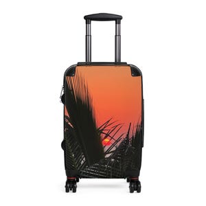 Pode incluir: Uma mala preta com rodas com um design de pôr do sol. A imagem na mala mostra um céu laranja e vermelho vibrante com o sol se pondo atrás de folhas de palmeira verde escuras. A mala tem uma alça retrátil.