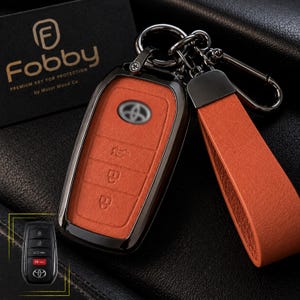 May include: An orange leather key fob cover for a Toyota vehicle, featuring a metallic frame, a matching orange leather strap, and a dark metal keyring. The cover includes embossed button icons and a Toyota logo cutout. A small inset shows the original black key.