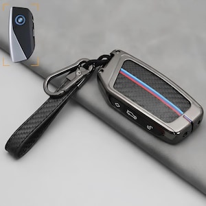May include: A dark gray car key fob cover featuring a carbon fiber texture inlay with a diagonal blue and red stripe. The case includes a matching carbon fiber patterned strap and a black metal carabiner clip for secure attachment to keys.