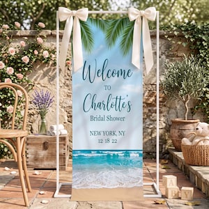May include: A vertical rectangular banner featuring a beach scene with turquoise ocean waves and sand. The text reads Welcome to Charlotte's Bridal Shower, New York, NY, 12 18 22. The banner is held by a white metal frame with cream bows.