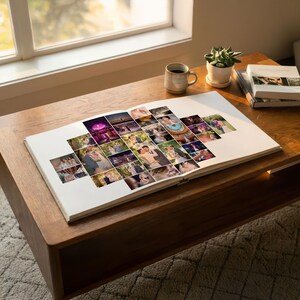 May include: A white photo album lies open, displaying a collage of various wedding photographs arranged in a central cluster on the page. The album is positioned on a wooden surface next to a ceramic mug and a stack of books near a window.