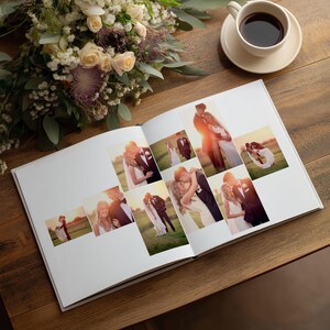 May include: An open photo album featuring a collection of wedding photographs displayed on white pages. The album is placed next to a bouquet of cream and purple flowers and a white ceramic cup filled with dark coffee on a wooden surface.