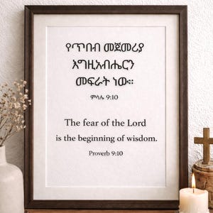 May include: A framed print with a dark brown frame displays the text "The fear of the Lord is the beginning of wisdom. Proverb 9:10." A lit candle, a small cross, and a vase with flowers are on a wooden surface.