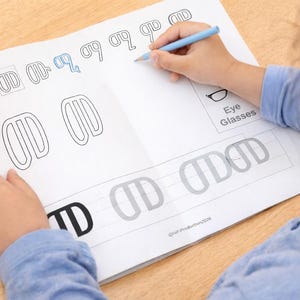 Peut inclure: Un enfant trace des lettres sur une feuille de travail avec un crayon bleu. La feuille de travail présente des contours de lettres et les mots "Eye Glasses". Une tasse rouge avec des crayons de couleur est à gauche. Le livre est intitulé "First Coloring and Tracing Book".