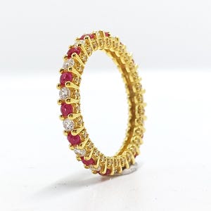 May include: A gold-colored eternity ring with alternating red and clear gemstones. The ring is set on a white surface, showcasing the vibrant colors of the gemstones. The ring's design is a continuous band of stones, creating a classic and elegant look.