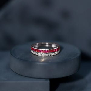 Peut inclure: Une bague en argent ornée d'une rangée de petits rubis carrés et d'une rangée de petits diamants clairs. La bague est mise en valeur sur un fond bleu foncé, soulignant l'éclat des pierres. Un bijou au design classique.