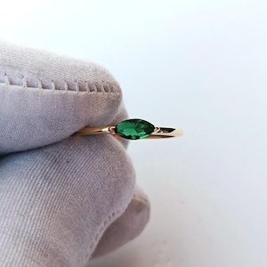 Puede incluir: Un anillo de oro con una gema verde esmeralda de talla marquesa. El anillo tiene una banda delgada y dorada. La gema está sujeta por pequeñas garras. El anillo está sostenido por una mano enguantada.