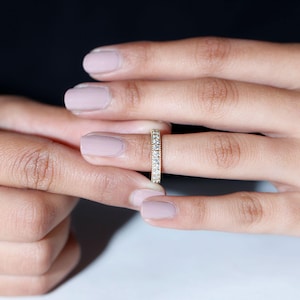 Peut inclure: Une bague en or avec une rangée continue de petites pierres précieuses claires. La bague est tenue entre deux doigts, mettant en valeur son design délicat et son éclat. Un bijou classique.