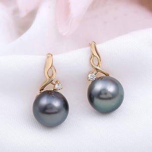 May include: A pair of gold-toned drop earrings featuring large, dark gray pearls. Each earring has a swirling gold design with a small, clear gemstone accent. The earrings are displayed on a white, textured surface.