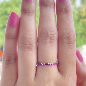 May include: A close-up of a hand wearing a thin, silver ring. The ring is adorned with small, rectangular, pink gemstones. The person's nails are painted a bright pink color, complementing the ring's gemstones. The background is blurred, with hints of green.