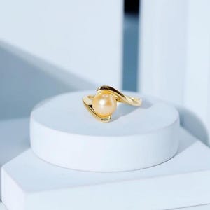 Könnte beinhalten: Ein goldfarbener Ring mit einer großen, runden, hellorangenen Perle. Das Band des Rings hat ein fließendes, asymmetrisches Design. Der Ring wird auf einer weißen, geometrischen Oberfläche vor hellblauem Hintergrund präsentiert.