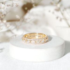 Pode incluir: Um anel de ouro com três fileiras de pedras preciosas brilhantes. O anel está sobre um pedestal branco e redondo, com detalhes suaves e desfocados no fundo. As pedras são redondas e em forma de marquesa.