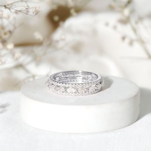 Könnte beinhalten: Ein silberner Ring mit mehreren Reihen kleiner, klarer Edelsteine. Der Ring liegt auf einem weißen, kreisförmigen Sockel, mit weichen, verschwommenen floralen Elementen im Hintergrund. Das Design des Rings kombiniert runde und marquiseförmige Steine.