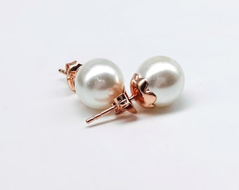 May include: A pair of pearl stud earrings with rose gold-colored floral accents. The earrings feature large, round, white pearls and are set on posts with butterfly backs. The earrings are displayed on a white background.