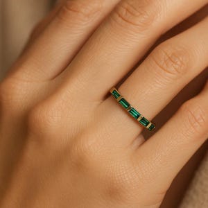 May include: A gold ring featuring a row of emerald-cut green gemstones. The ring is displayed on a finger, highlighting the design and colour of the jewellery. The ring's band is a thin, polished gold.