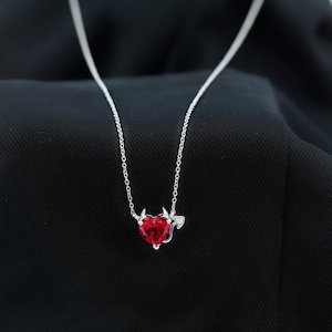 May include: A silver necklace featuring a red heart-shaped pendant with devil horns and a tail. The pendant is made of a shiny, red gemstone. The necklace chain is delicate and silver.