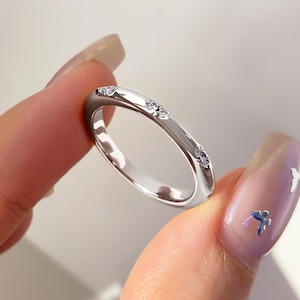 Puede incluir: Un anillo plateado con tres pequeñas piedras preciosas transparentes engastadas a lo largo de la banda. El anillo tiene un acabado liso y pulido y un diseño ligeramente curvado. El anillo se sostiene entre dos dedos.