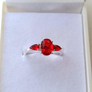 Peut inclure: Une bague en argent ornée d'une grande pierre rouge ovale flanquée de deux petites pierres rouges en forme de poire. La bague est présentée dans un écrin blanc, mettant en valeur la couleur vive et le design du bijou.