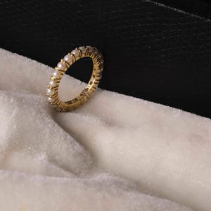 Puede incluir: Un anillo de oro con una banda de pequeñas perlas redondas. El anillo se presenta sobre una tela blanca texturizada y un fondo negro. Una joya atemporal, adecuada para diversas ocasiones.