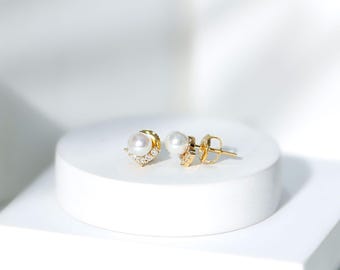 May include: A pair of gold stud earrings with a pearl center and small, clear gemstones. One earring has a decorative, v-shaped design. The earrings are displayed on a white, round surface.