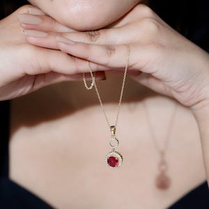 Könnte beinhalten: Eine goldfarbene Halskette mit einem Anhänger, der einen großen, runden, roten Edelstein umgeben von kleineren, klaren Steinen aufweist. Der Anhänger hängt an einer zarten Kette. Die Halskette wird von den Händen einer Person gehalten.