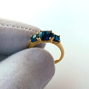 Op de afbeelding: Een gouden ring met drie donkerblauwe edelstenen. De band van de ring is goudkleurig en de edelstenen zijn gezet in sierlijke, goudkleurige zettingen. De ring wordt tegen een witte achtergrond gehouden.