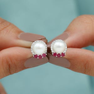 May include: A pair of pearl stud earrings. Each earring features a large, round, white pearl surrounded by a halo of small, clear gemstones. Below the pearl are three small, red gemstones. The earrings are set in a silver metal.