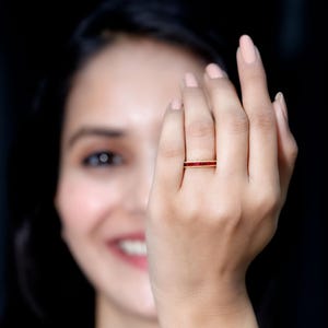Könnte beinhalten: Ein goldener Bandring mit einer Reihe kleiner, roter Edelsteine. Der Ring befindet sich am Finger einer Person, wobei das Gesicht der Person teilweise im Hintergrund zu sehen ist. Der Ring ist ein zarter Schmuck.