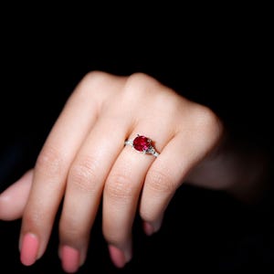 May include: A silver ring with a large, oval-cut, deep red gemstone. Two smaller, clear gemstones are either side of the main stone. The ring is set against a dark background, accentuating the vibrant colour of the central stone.