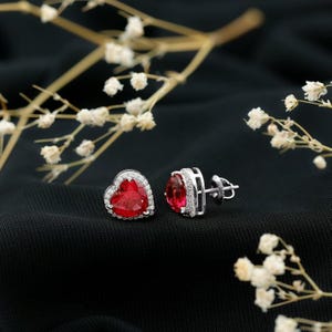 May include: A pair of heart-shaped stud earrings with a vibrant red center stone. The earrings are framed by a halo of small, clear stones and set in a silver-colored metal. The background is black, with white floral accents.