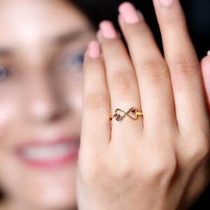 Pode incluir: Um anel dourado em forma de símbolo de infinito, com dois detalhes em forma de coração, cada um contendo uma pequena pedra preciosa vermelha. O anel é exibido em um dedo, com um fundo desfocado.