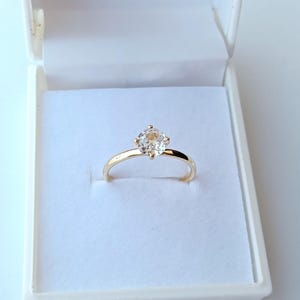 Puede incluir: Un anillo de oro con una gema grande y transparente de talla cuadrada. El anillo está engastado en un estilo sencillo y clásico y se muestra dentro de una caja de joyería blanca.