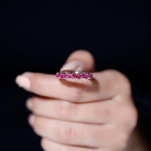 May include: A silver ring with a band of vibrant pink gemstones. The ring is held up against a dark background, showcasing the sparkle of the gemstones. The ring is a piece of jewelry.