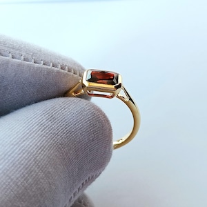 Könnte beinhalten: Ein goldener Ring mit einem rechteckigen, rotbraunen Edelstein. Der Ring hat ein schlichtes Design mit einem dünnen Band und einer Lünettenfassung. Die Form und Farbe des Edelsteins werden hervorgehoben. Heller Hintergrund.