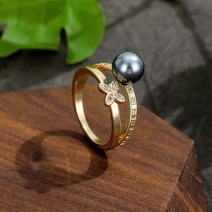 Può includere: Anello color oro con una perla grigio scuro e un motivo floreale con piccole pietre trasparenti. L'anello ha una doppia fascia ed è appoggiato su una superficie di legno.