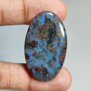 Könnte beinhalten: Ovaler Edelstein mit blauer Grundfarbe, gesprenkelt mit braunen, weinroten und grünen Einschlüssen. Der polierte Stein wird in einer Hand gehalten, wodurch seine natürlichen Muster hervorgehoben werden. Ein einzigartiger Stein für Schmuck oder Sammlungen. Ca. 4 cm.