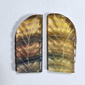 May include: Two semi-circular, polished gemstones with carved leaf patterns. The stones display a mix of yellow, brown, and purple hues. The leaf veins are etched into the surface, creating a textured effect. The gemstones are set against a white background.